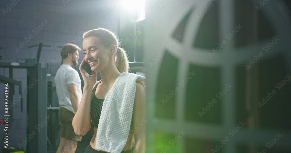 Cheerful woman is standing at the gym and holding smartphone while enjoying of the pleasure chat with somebody. She is having break between work out in exercising room Cheerful woman is standing at the gym and holding smartphone while enjoying of the pleasure chat with somebody. She is having break between work out in exercising room