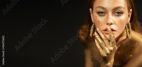 Stylish girl looks at camera. Gold ring on finger, earrings rings. Beautiful face, blue eyes, blonde hair golden glamorous makeup, gold paint on body, skin, hands. Portrait of fashion model woman