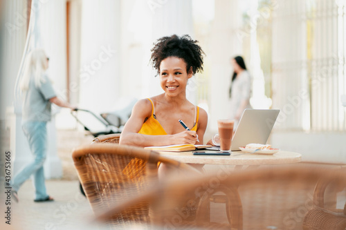 A student in a cafe, a young woman working smiling typing a message on a laptop. African-American appearance and black curly hair. Sitting in a summer restaurant after a pandemic.