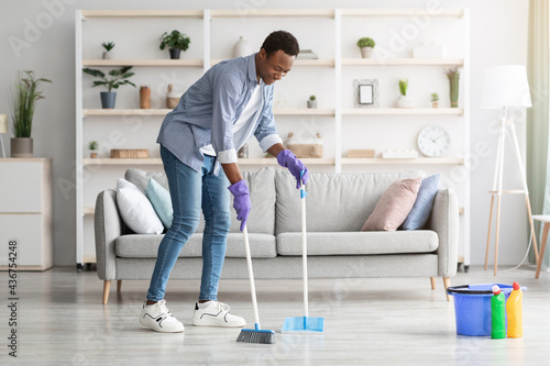 Cheerful black guy cleaning living room, holding broom