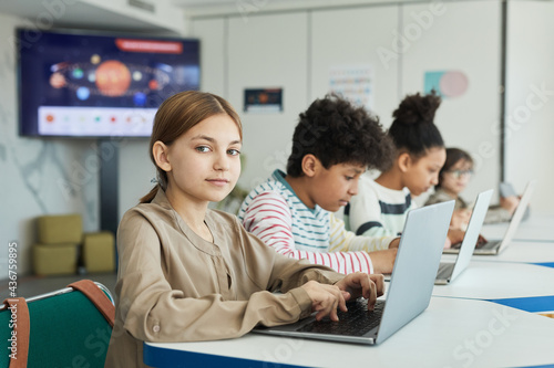 Portrait of young schoolgirl looking at camera while using laptop in IT class  copy space