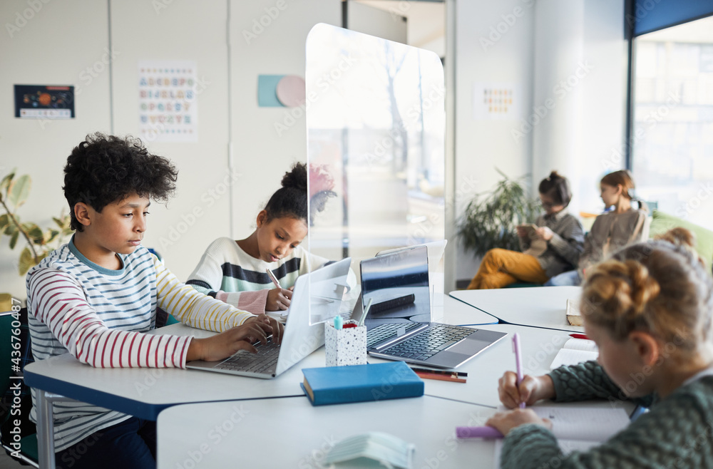 Obraz premium Diverse group of children working together at desk in classroom with protective glass separations, copy space