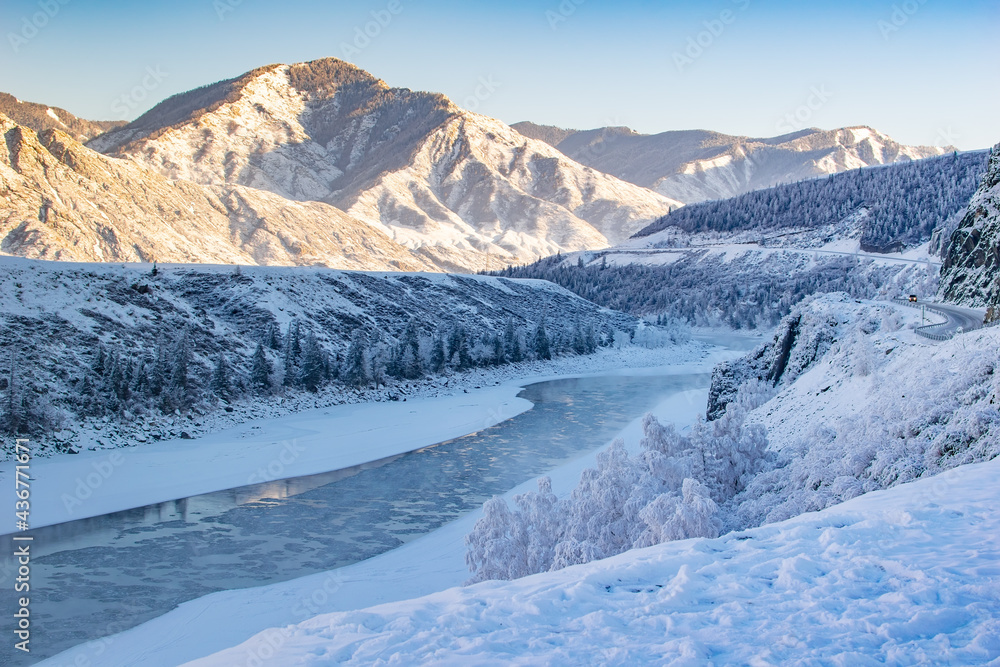 winter, frosty, snowy landscape of the valley with a river, high mountain peaks with sharp bare stones, a winding highway and passing cars on it and clear sunny weather