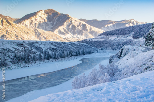 winter, frosty, snowy landscape of the valley with a river, high mountain peaks with sharp bare stones, a winding highway and passing cars on it and clear sunny weather