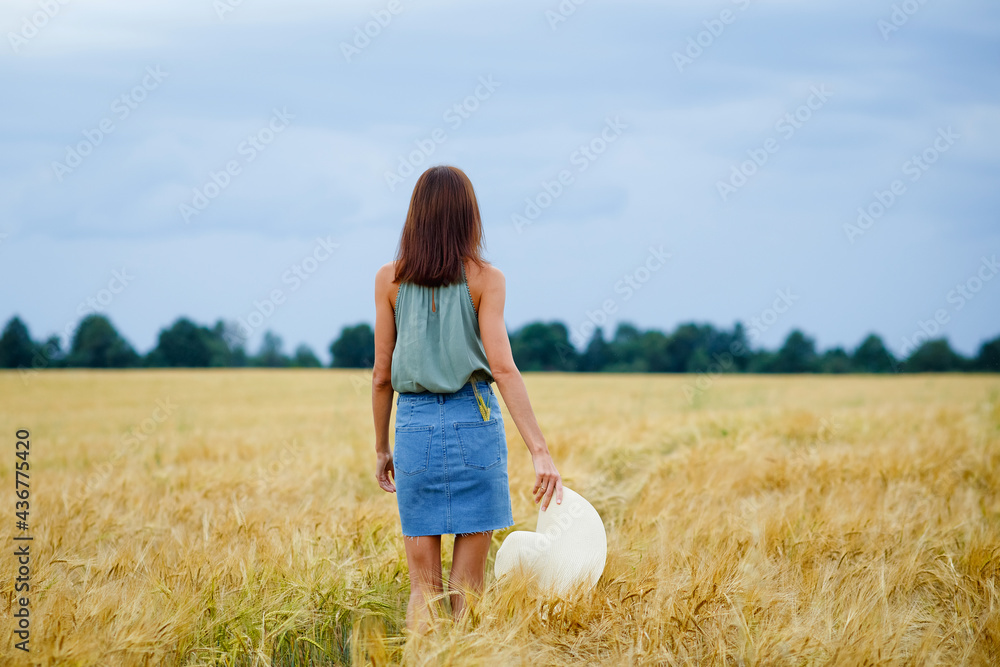 A young woman with brown hair in a denim skirt walks through a wheat field, holding a hat in her hand. View from the back. Rural landscape. Summer mood. A young woman with brown hair in a denim skirt walks through a wheat field, holding a hat in her hand. View from the back. Rural landscape. Summer mood.