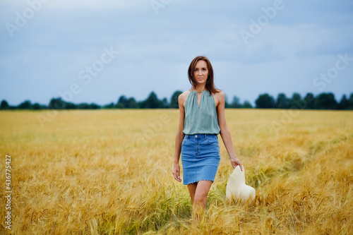 Fototapeta Naklejka Na Ścianę i Meble -  A young woman with brown hair in a skirt walks through a wheat field, holding a hat in her hand. Rural landscape. Summer mood.