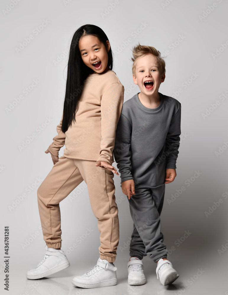 Fototapeta premium Kids in cosy cotton sportswear play together. Happy blond kid boy and brunette kid girl in pants and hoodies stands close to each other screaming and laughing over gray background