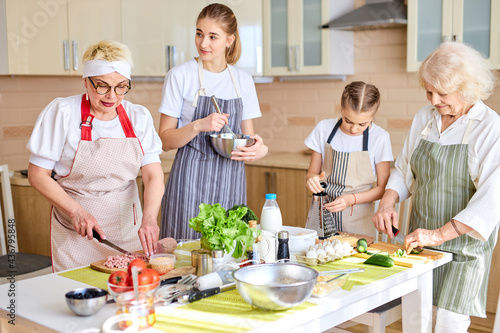 friendly different generation family preparing meal for dinner together, talking, good time in domestic atmosphere, dressed in apron, at home in cozy bright kitchen