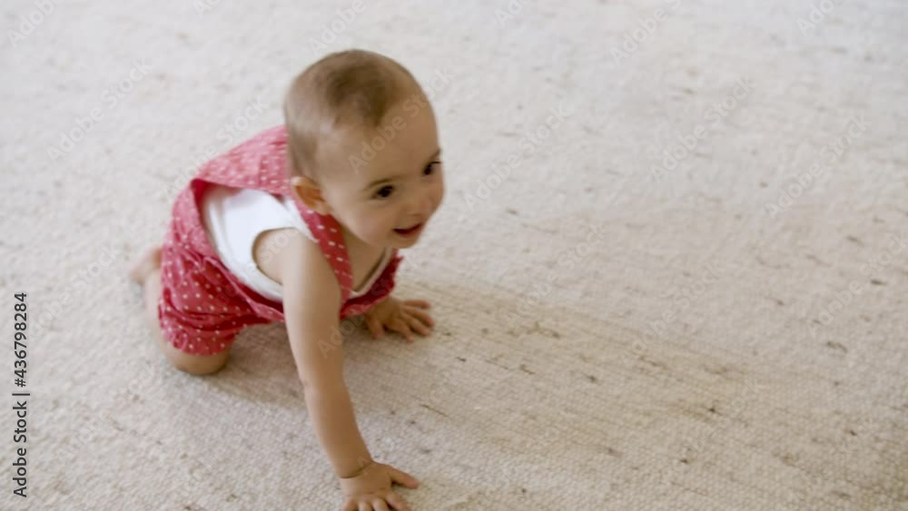 Pretty kid crawling on carpet in living room. Happy girl in red dungarees learning how to walk, sitting on floor, trying to stand up. Childhood, family concept. Pretty kid crawling on carpet in living room. Happy girl in red dungarees learning how to walk, sitting on floor, trying to stand up. Childhood, family concept.