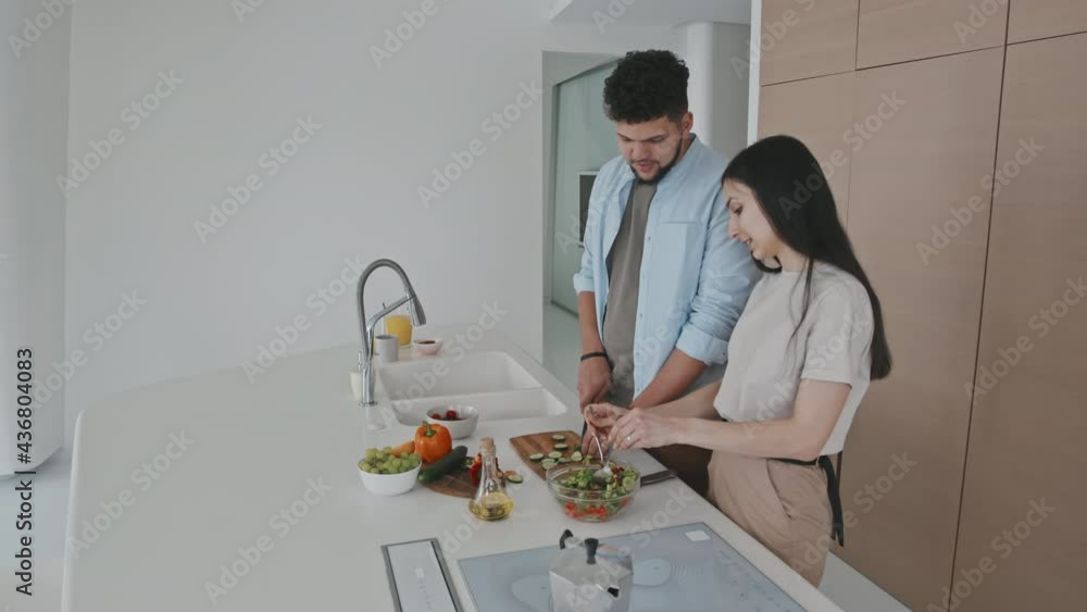Medium shot of handsome young man of Latin ethnicity helping his attractive girlfriend to cut vegetables for salad standing together at modern minimalist kitchen and chatting Medium shot of handsome young man of Latin ethnicity helping his attractive girlfriend to cut vegetables for salad standing together at modern minimalist kitchen and chatting