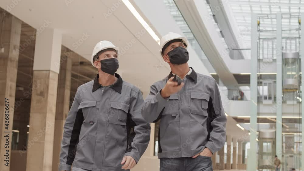 Medium tracking of two Caucasian men wearing white hard hats and facial masks, walking in large building under construction, looking around Medium tracking of two Caucasian men wearing white hard hats and facial masks, walking in large building under construction, looking around