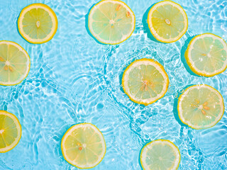 Lemon slices in clean transparent water over blue background with copy space. Water splashing on blue water surface in sunlight. Top view or flatlay. Summer, vacation, healthy eating concept