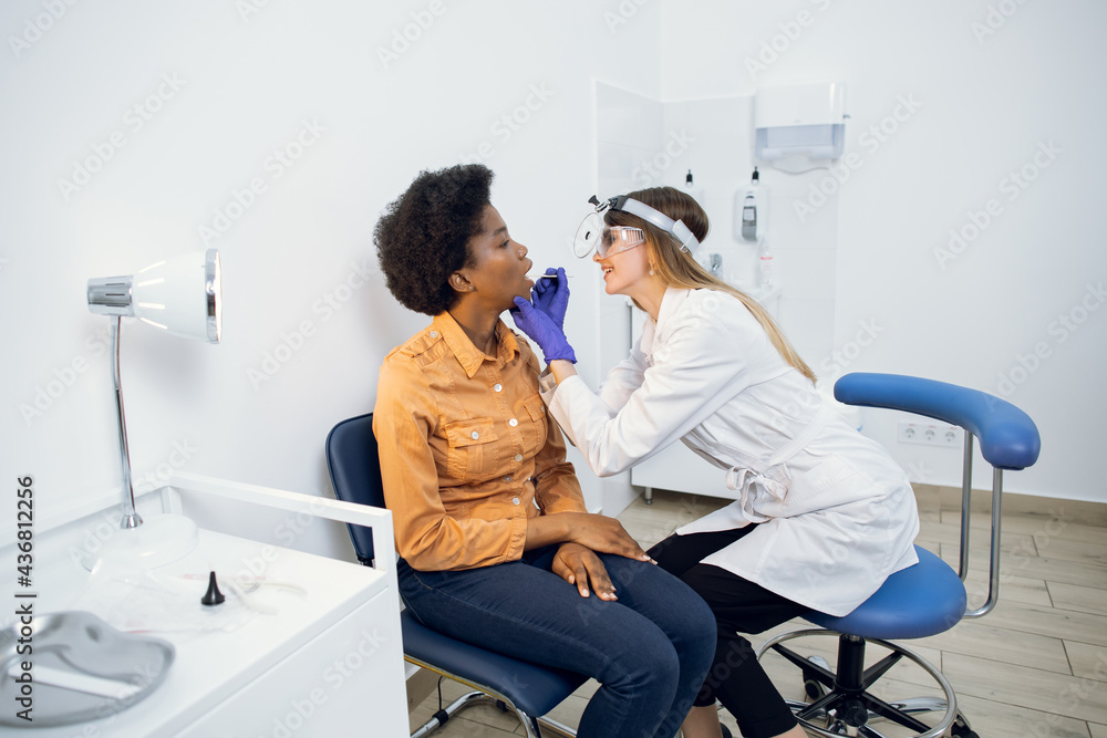 Fototapeta premium Pharyngitis and laryngitis treatment concept. Professional female doctor laryngologist, looking at throat of african american woman patient with wooden spatula in modern clinic