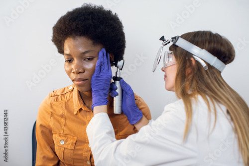 Ear deseases, otitis, hearing loss, otolaryngology concept. Caucasian professional ent doctor inspecting the ear canal and eardrum of young African woman patient with otoscope