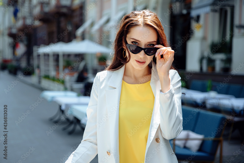 Fashion portrait of attractive woman in white jacket with long hair. Charming lady in sunglasses looking at camera. Happy stylish girl in city street