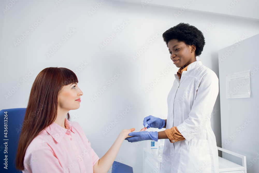 Female African American lab technician, drawing finger blood for test. Young Caucasian lady patient trusting her doctor and having blood test