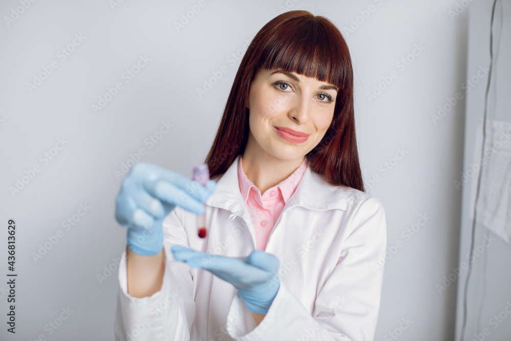 Close up portrait of young pleasant Caucasian lady doctor, lab technician, researcher, holding in hands test tube with blood sample, posing to camera in modern light medical lab or clinic Close up portrait of young pleasant Caucasian lady doctor, lab technician, researcher, holding in hands test tube with blood sample, posing to camera in modern light medical lab or clinic