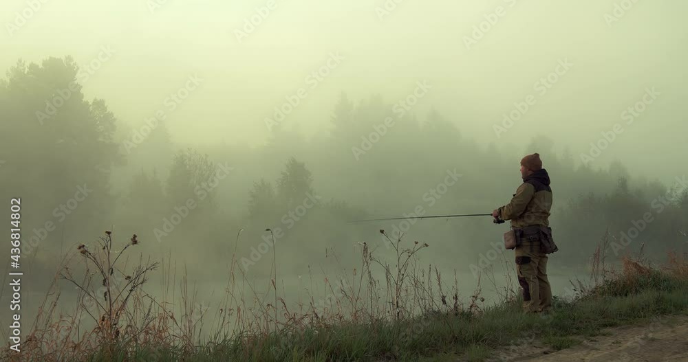 A man in a special warm suit for protection against insects throws a spinning rod, turns the reel and lures the fish. There is a thick fog over the lake, a forest can be seen behind the reservoir A man in a special warm suit for protection against insects throws a spinning rod, turns the reel and lures the fish. There is a thick fog over the lake, a forest can be seen behind the reservoir