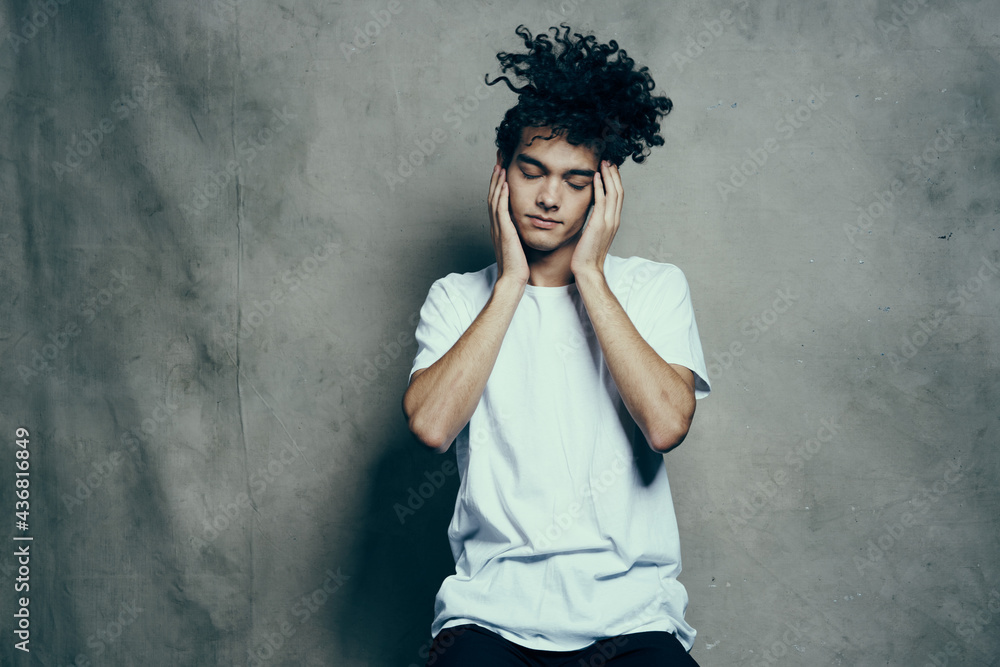 Cheerful guy with curly hair gestures with his hands near his face on a gray fabric background in a white t-shirt