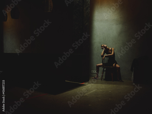 a woman spread her legs sits on a chair In a dark room and looks at the light from above