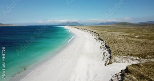 4k aerial footage of West Beach, Berneray, Outer Hebrides, Scotland. Sunny day, white sand, turquoise and blue sea water, sand dunes with green grass, blue sky and light cloud, mountains in distance.