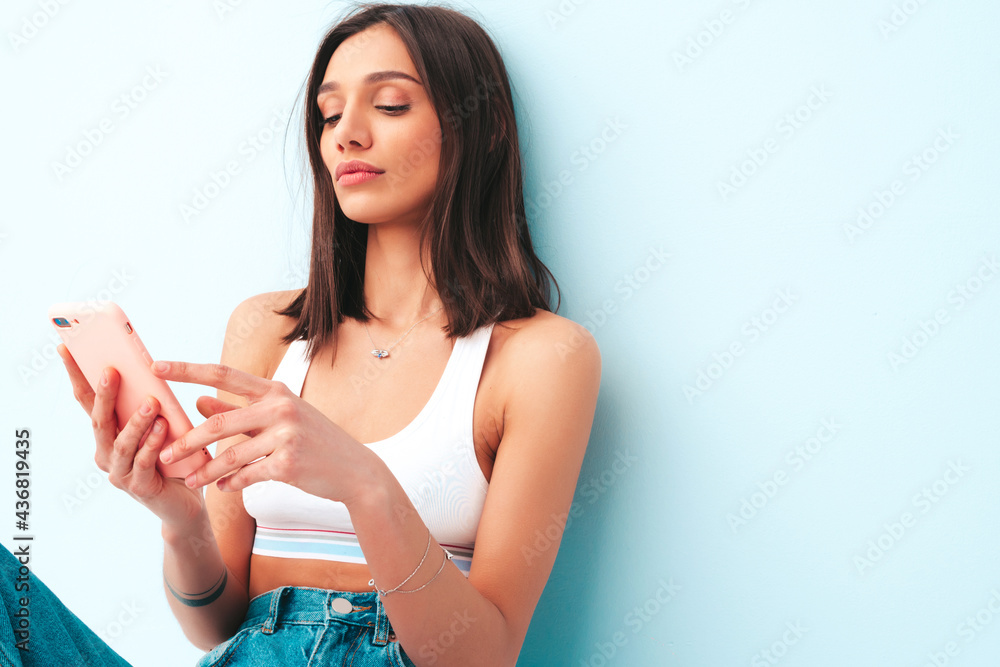 Beautiful smiling woman dressed in white jersey top shirt and jeans. Sexy carefree cheerful model enjoying her morning. Positive female posing near light blue wall in studio.Using mobile apps Beautiful smiling woman dressed in white jersey top shirt and jeans. Sexy carefree cheerful model enjoying her morning. Positive female posing near light blue wall in studio.Using mobile apps