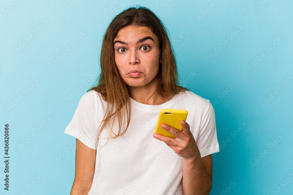 Young caucasian woman holding a mobile phone isolated on blue background shrugs shoulders and open eyes confused. Young caucasian woman holding a mobile phone isolated on blue background shrugs shoulders and open eyes confused.
