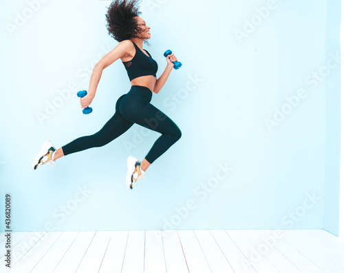 Fitness smiling black woman in sports clothing with afro curls hairstyle.She running and jogging . Young beautiful model with perfect tanned body.Female jumping with dumbbells in studio near blue wall