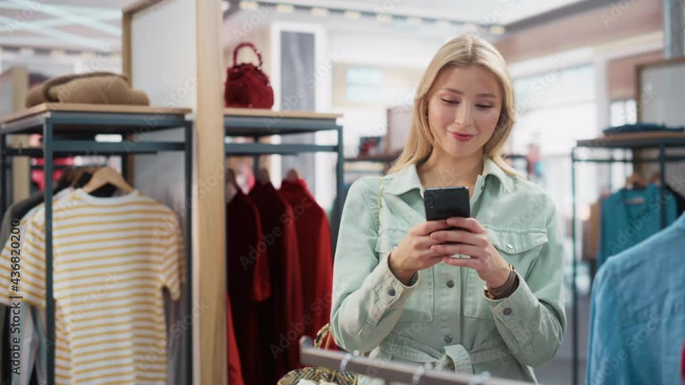Beautiful Smiling Female Customer Shopping in Clothing Store, Using Smartphone, Browsing Online, Comparing on Internet, Choosing Stylish Clothes. Fashionable Shop, Colorful Brands, Sustainable Designs Beautiful Smiling Female Customer Shopping in Clothing Store, Using Smartphone, Browsing Online, Comparing on Internet, Choosing Stylish Clothes. Fashionable Shop, Colorful Brands, Sustainable Designs