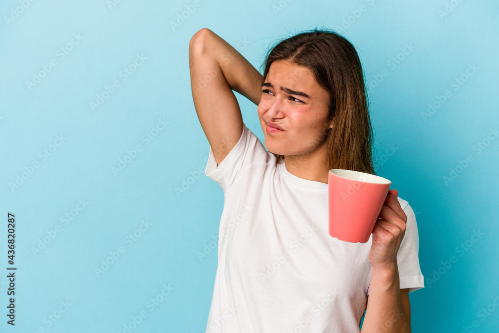 Young caucasian woman holding a mug isolated on blue background touching back of head, thinking and making a choice. Young caucasian woman holding a mug isolated on blue background touching back of head, thinking and making a choice.