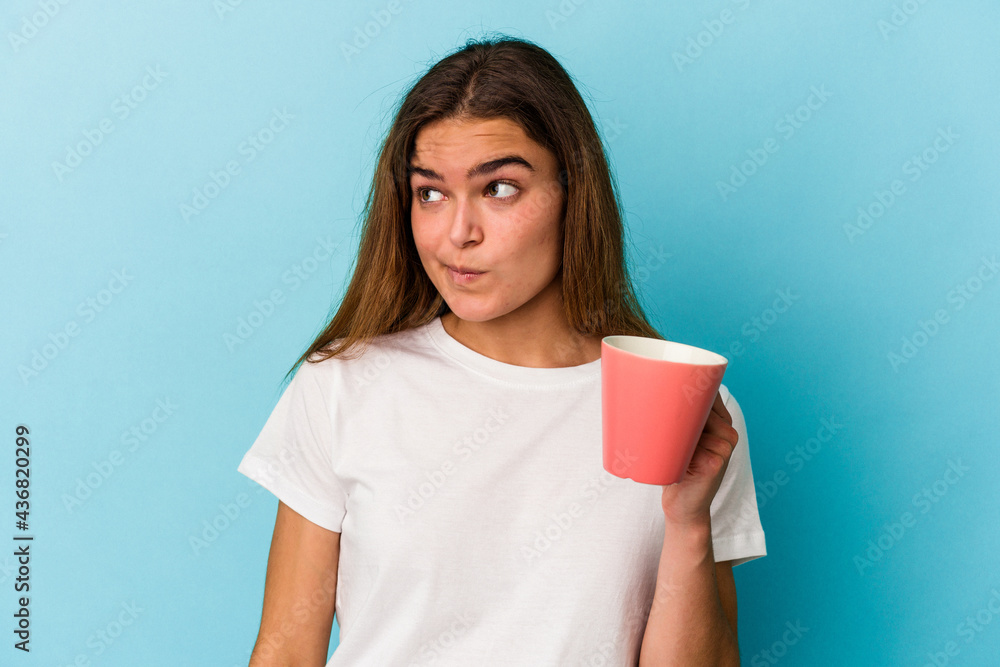 Young caucasian woman holding a mug isolated on blue background confused, feels doubtful and unsure. Young caucasian woman holding a mug isolated on blue background confused, feels doubtful and unsure.