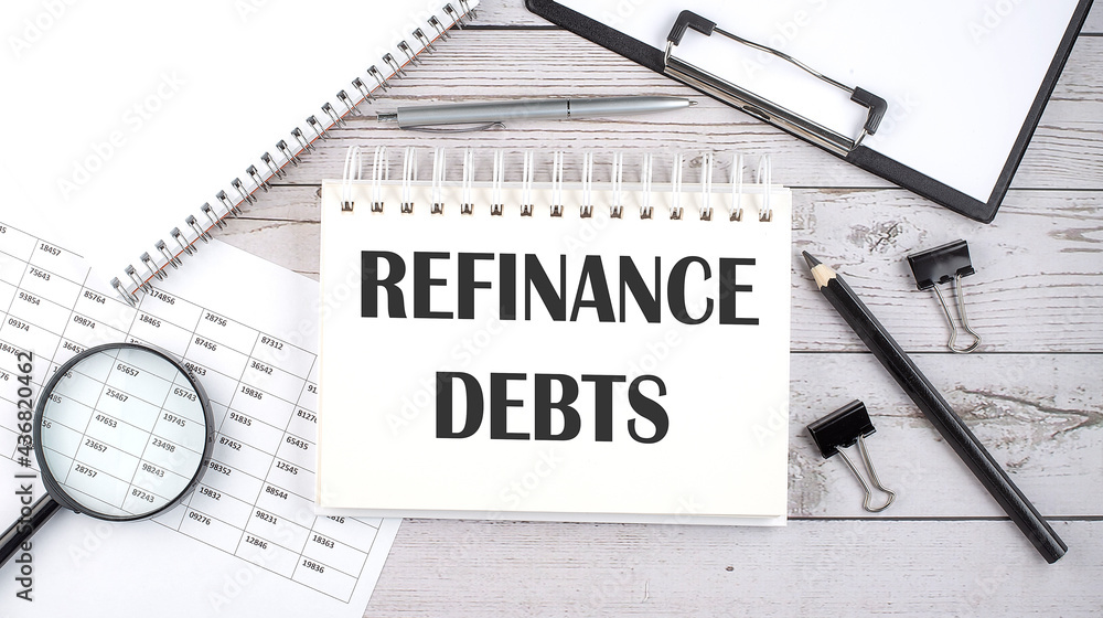 Notepad with text REFINANCE DEBTS with a pen,office tools and graphs on the desktop. Notepad with text REFINANCE DEBTS with a pen,office tools and graphs on the desktop.