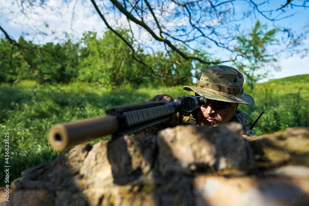 A soldier in camouflage uniforms aiming at the scope with a weapon from behind cover