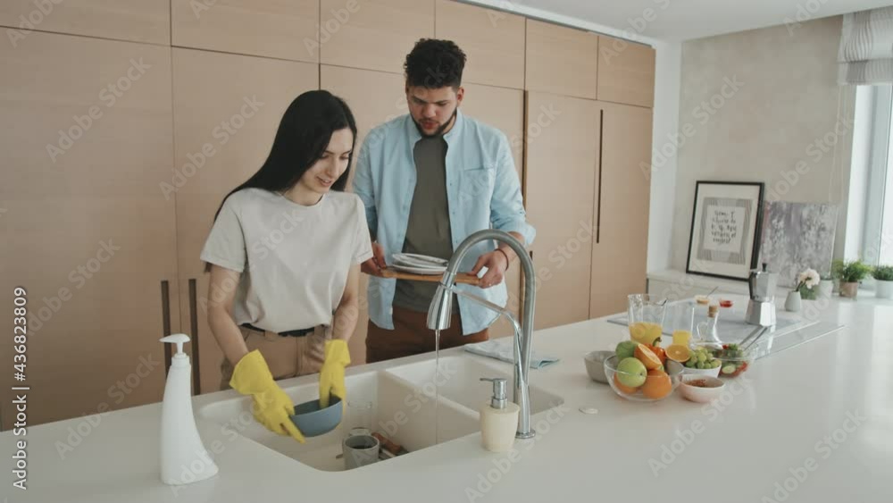 Medium shot of happy young Latin man helping his beautiful wife to dry dishes while she washing them standing at modern design kitchen in minimalist style Medium shot of happy young Latin man helping his beautiful wife to dry dishes while she washing them standing at modern design kitchen in minimalist style