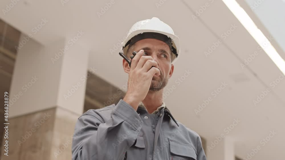 Low angle of blue-eyed Caucasian construction worker wearing white safety helmet, communicating via walkie-talkie in mall under construction Low angle of blue-eyed Caucasian construction worker wearing white safety helmet, communicating via walkie-talkie in mall under construction