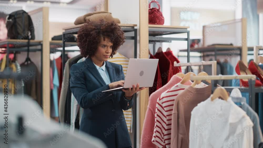 Clothing Store: Female Merchandising Manager Uses Laptop Computer To Create Stylish Fashion Collection. Professional Shop Sales Retail Assistant Checks Stock. Small Business Owner Orders Mall Items. Clothing Store: Female Merchandising Manager Uses Laptop Computer To Create Stylish Fashion Collection. Professional Shop Sales Retail Assistant Checks Stock. Small Business Owner Orders Mall Items.