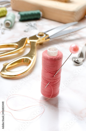 Closeup of pink spool of thread, sewing pin, golden scissors and other tailor tools on the table