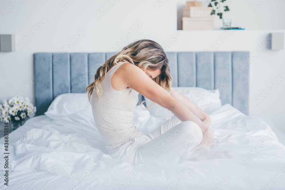 Long haired blonde in pyjama wakes and stretches back sitting on large white bed with blurred flowers on foreground in early morning at home. Long haired blonde in pyjama wakes and stretches back sitting on large white bed with blurred flowers on foreground in early morning at home.