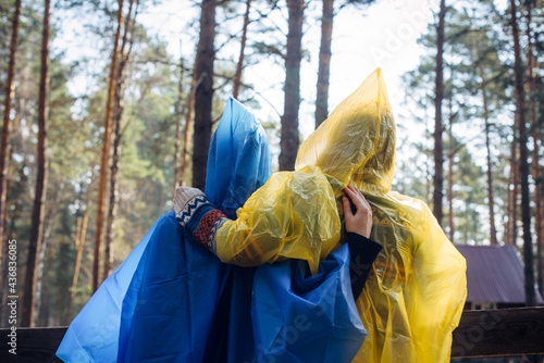 Two friends in hooded raincoats standing back to the camera in a tourist parking lot in the woods. Women in colorful raincoats rest during a hike. Camping concept.
