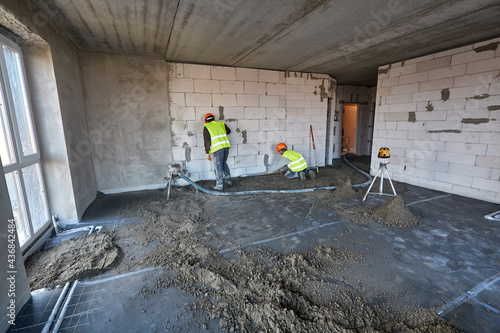 Two workers using pneumatic conveyor and tacheometer in an apartment of building under construction. Preparing for making a cement floor screed