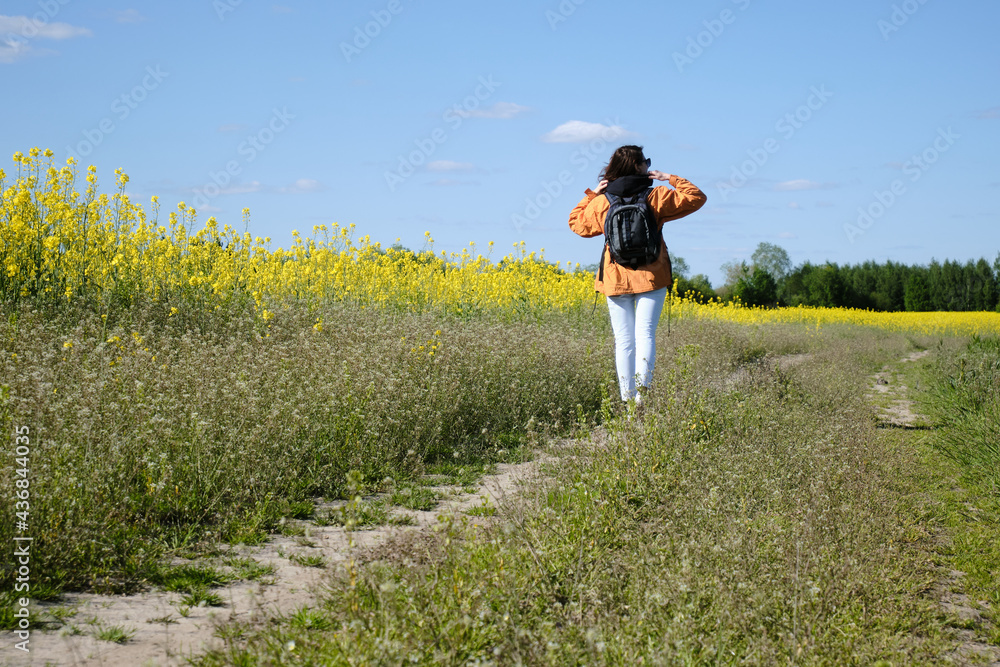 A girl walks along a rural road along a meadow with flowers. Travel outside the city. Sunny weather. Meadow with flowers