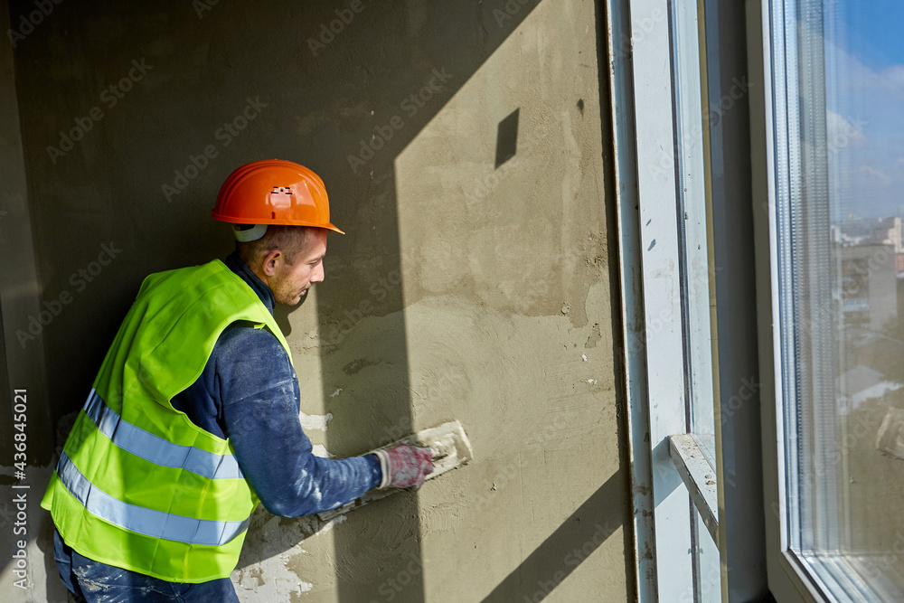 Worker in workwear and safety helmet is putting putty on wall, using spatula. Apartment of building under construction with panoramic windows Worker in workwear and safety helmet is putting putty on wall, using spatula. Apartment of building under construction with panoramic windows
