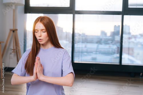 Close-up of redhead focused young woman meditating sitting on floor in lotus pose, Namaste mudra in light office room. Calm pretty redhead lady relaxation during yoga workout at home with closed eyes.