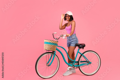 Full length portrait of emotional black lady in summer clothes standing with bike on pink studio background, copy space © Prostock-studio
