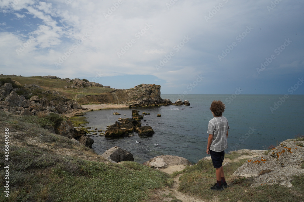A teenage boy stands on the seashore and looks into the distance. Teenager on the rocky seashore alone.