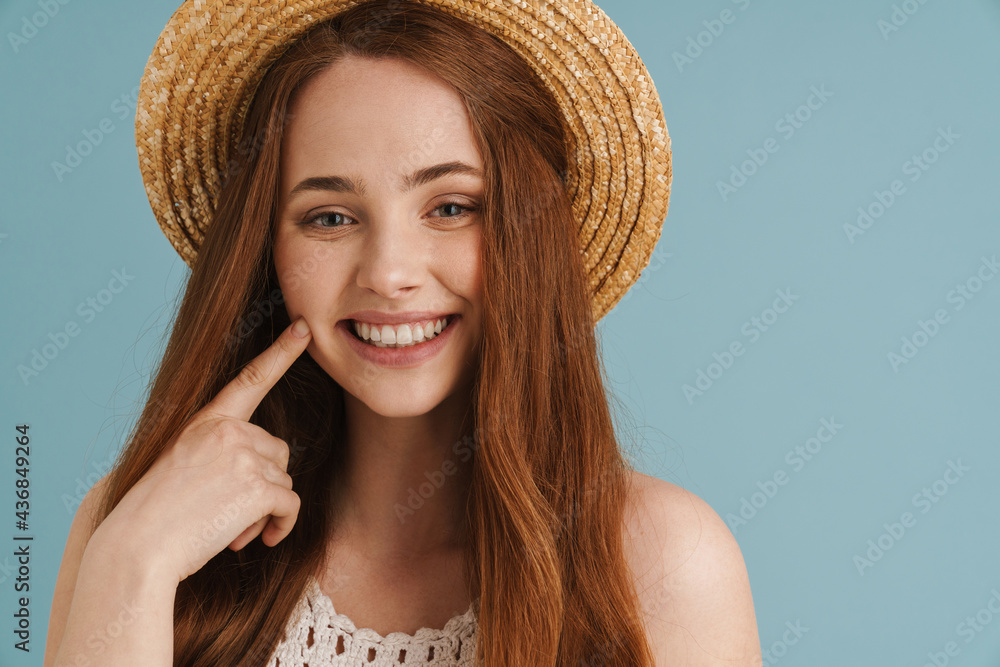 Young ginger woman in straw hat smiling and pointing finger at her cheek