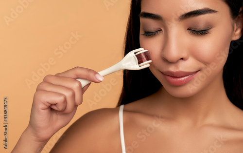 Close-up of Asian female face with dermaroller for mesotherapy procedures, skin care at home and in the salon. Mesoscooter with microneedles on a beige background. photo