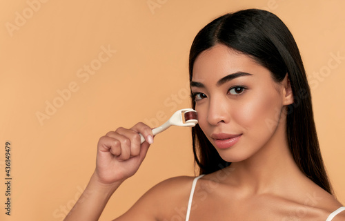 Close-up of Asian female face with dermaroller for mesotherapy procedures, skin care at home and in the salon. Mesoscooter with microneedles on a beige background. photo