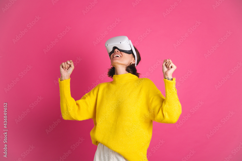 Beautiful woman in bright yellow sweater on pink background in virtual reality glasses happy jumping clenching fist winner gesture