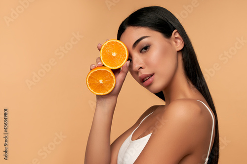 Portrait of Asian girl with shining clean skin of face holding orange halves in white underwear isolated on beige background. Vitamin C cosmetics concept photo